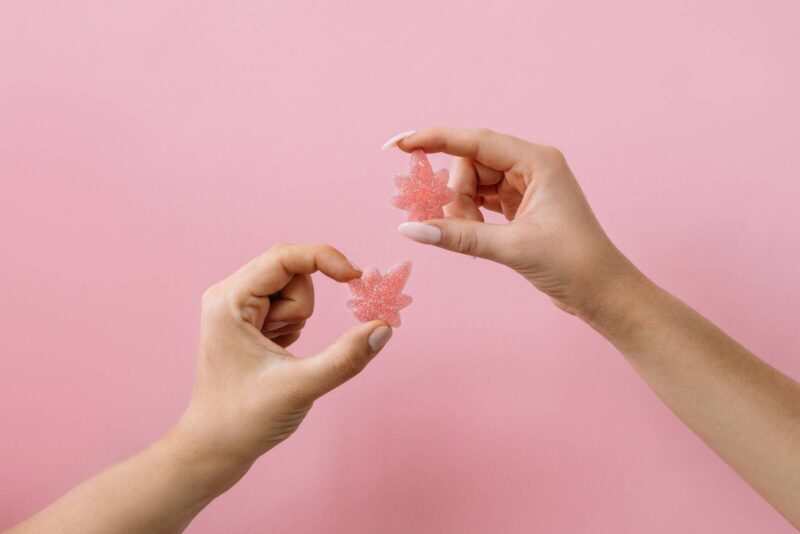 Two weed leaf-shaped pink gummies being held up in front of a pink background