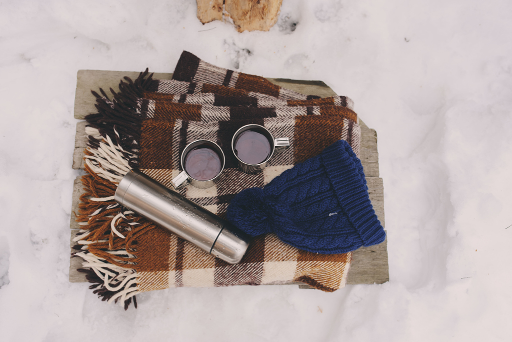 A blanket, thermos, two mugs and a toque laying in the snow