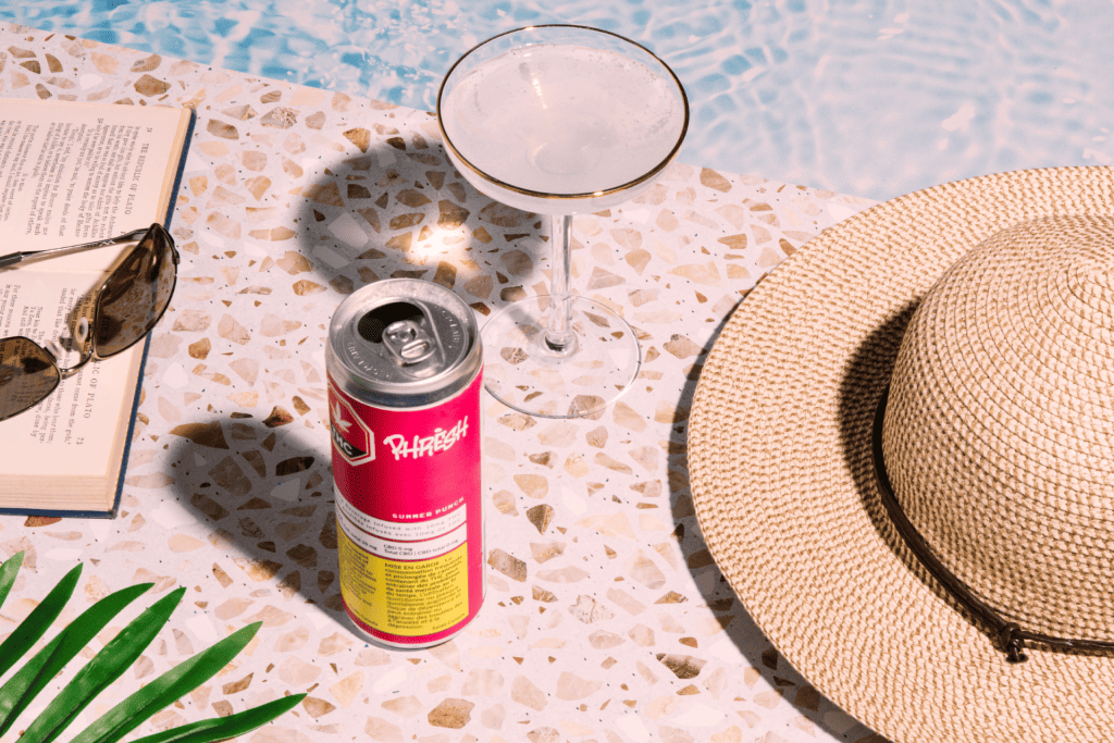  a cannabis-infused drink alongside a glass, hat, sunglasses, and book 